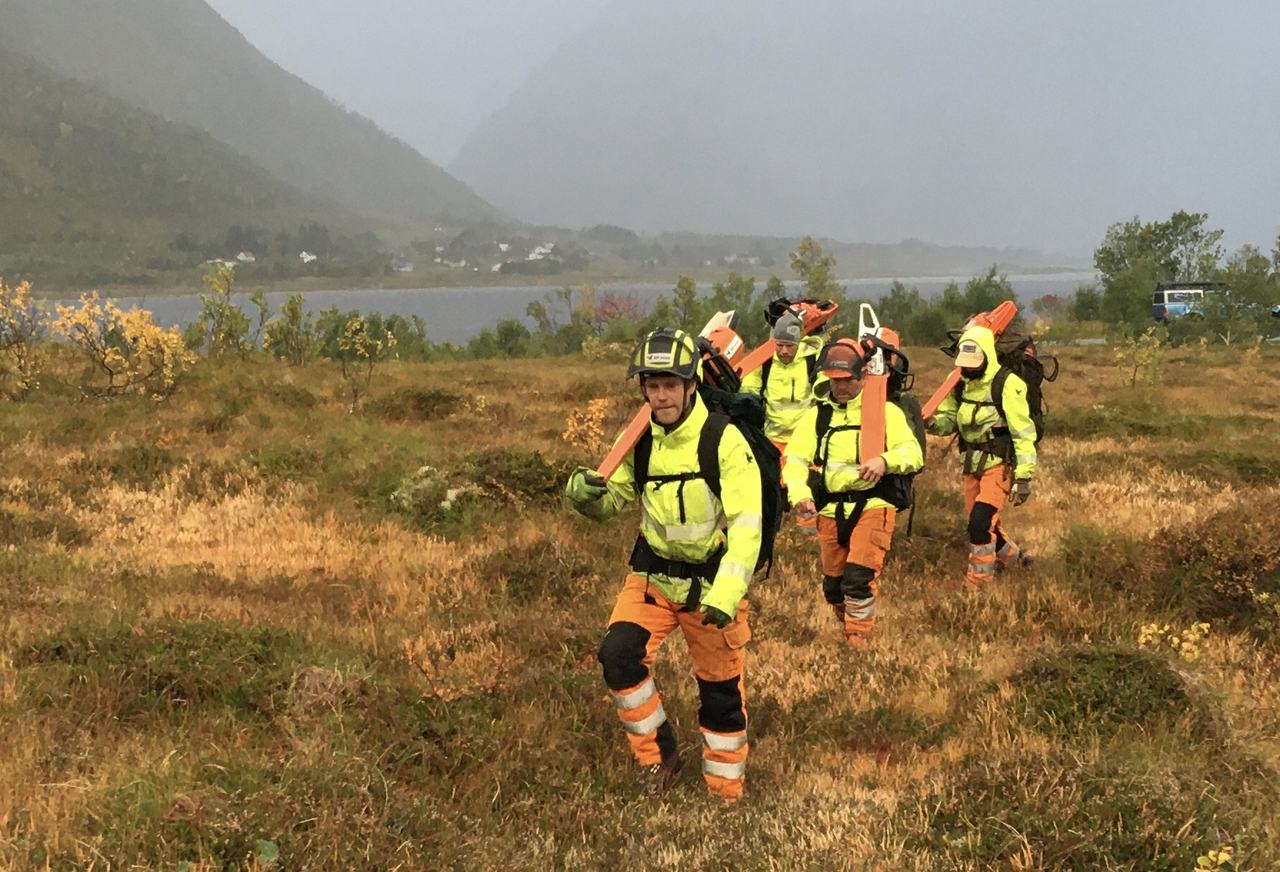 Fire arborister i refleksklær og hjelm går over lyngmyr i Lofoten, med fjell i skodde bak